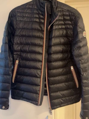 Svart dunjacka från Moncler - Snygg svart dunjacka från Moncler med quiltad design och hög krage. Jackan har dragkedja framtill med röd, vit och blå detalj samt två fickor med matchande dragkedjor. Moncler-logga på ärmen. Perfekt för kalla vinterdagar. Självklart äkta