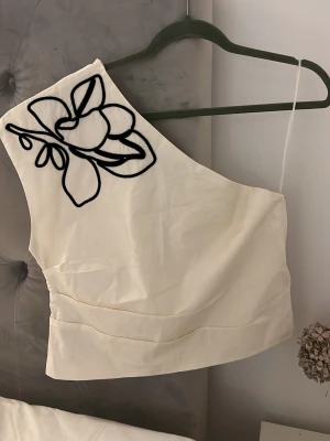 Vit  topp  -  Vit topp med asymmetrisk axel och svart broderat blommönster på ena sidan. Tunn axelrem på ena axeln och bredare axelparti på andra sidan. Diskret dragkedja på sidan. 