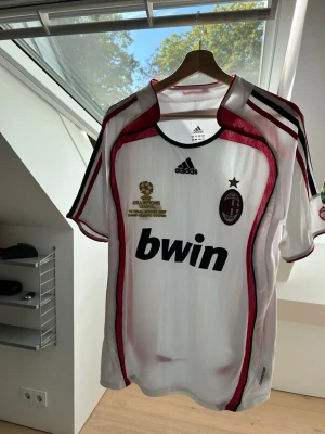 Retro AC Milan Adidas matchtröja - Säljer en vit AC Milan matchtröja från Adidas med röda och svarta detaljer. Tröjan har Champions League-märke, klubbmärke och Bwin-tryck på bröstet. Korta ärmar och klassisk sportig passform i lätt material, perfekt för fotboll eller träning. Storlek S men passar även M. 