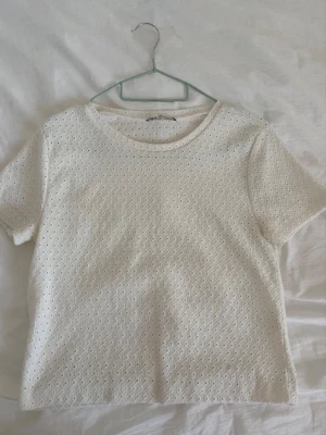 Vit kortärmad topp med broderade hål - Söt vit topp med korta ärmar och rund hals. Tyget har små broderade hål över hela plagget som ger en luftig och somrig känsla. Perfekt att matcha med jeans eller kjol för en fräsch look.