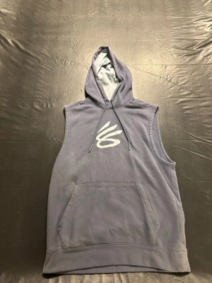 Ärmlös blå hoodie med tryck - Säljer en ärmlös hoodie i en snygg blå nyans med vit grafisk logga på bröstet. Hoodien har stor känguruficka framtill och justerbar huva med dragsko. Perfekt för en avslappnad och sportig stil.