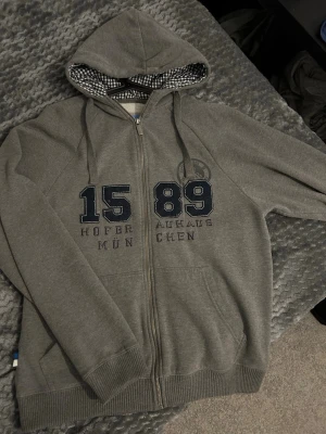 Grå HB München zip hoodie - Snygg grå zip hoodie från Hofbräuhaus München med tryck '1589' och text på bröstet. Huvan har rutigt innerfoder och dragsko. Mjuk insida och två fickor framtill. Perfekt för dig som gillar streetwear och vill ha något bekvämt och stilrent.