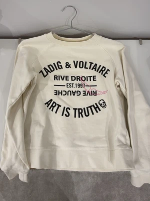 Vit sweatshirt Zadig & Voltaire - Snygg vit sweatshirt från Zadig & Voltaire med svart tryck framtill där det står 'ART IS TRUTH' och 'RIVE DROITE'. Tröjan har rund halsringning och en avslappnad passform. Perfekt för en cool och avslappnad stil.