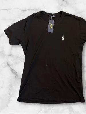 Svart t-shirt från Polo Ralph Lauren - Klassisk svart t-shirt från Polo Ralph Lauren med den ikoniska vita logobrodyr på bröstet. T-shirten har rund halsringning och korta ärmar. Tillverkad i mjuk bomull för en skön och avslappnad känsla.