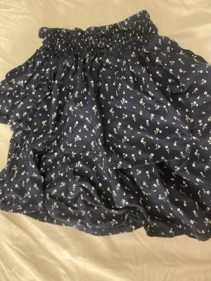 Mörkblå blommig volangkjol - Supersöt mörkblå kortkjol med små vita blommor och volangdetaljer. Kjolen har en bred resår i midjan som ger en bekväm passform och ett flowy fall. Perfekt för dig som gillar en feminin och trendig stil.