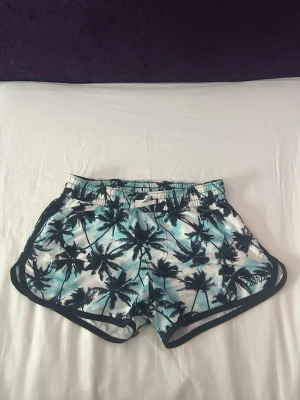 Badshorts - Säljer ett par snygga shorts med svart-vitt palmmönster och turkosa inslag. Shortsen har elastisk midja med vit snörning och svarta kanter. Perfekta för sommaren och strandhäng!