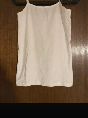 Vit linnetopp med smala axelband - Enkel och stilren vit linnetopp med smala axelband. Toppen har rak passform och ärmlös design, perfekt för varma dagar eller som lager under en skjorta. Materialet känns lätt och svalt.