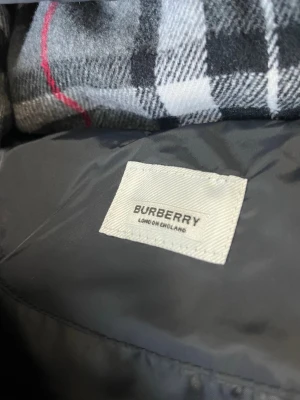 Svart pufferjacka från Burberry - Snygg svart pufferjacka från Burberry med klassiskt rutigt foder i huvan. Jackan har dragkedja, tryckknappar och två stora fickor framtill. Perfekt för kalla dagar och har en modern, boxig passform. Ikonisk detalj med Burberry-logga på ärmen.