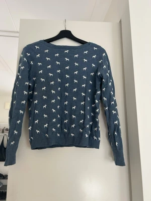 Blå stickad tröja med dalahästar - Supersöt blå stickad tröja från Jumperfabriken med vita små dalahästar som mönster över hela tröjan. Tröjan har rund halsringning, långa ärmar och ribbade muddar. Perfekt för dig som gillar lekfulla detaljer och vill ha något unikt i garderoben. Nypris ca. 2000kr.