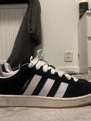 Adidas Campus svartvita sneakers - Mycket bra skicka. Klassiska Adidas Campus sneakers i svart mocka med vita läderdetaljer och de ikoniska tre ränderna på sidan. Skorna har vita skosnören och en låg silhuett med gummisula. Perfekta för en clean och stilren look.