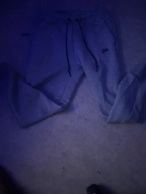 Gråa mjukisbyxor fleece från Nike  - Säljer ett par gråa mjukisbyxor från Nike med svarta dragsnören och logga på vänster ben. Byxorna har muddar vid bensluten och fickor med dragkedja. Perfekta för chill dagar eller träning.