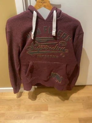 Vinröd hoodie från Superdry - Snygg vinröd hoodie från Superdry med tryck på bröstet och magficka. Hoodien har vita dragsnören och en klassisk huva. Perfekt för en avslappnad och trendig stil. Materialet är mjukt och skönt, passar bra till jeans.
