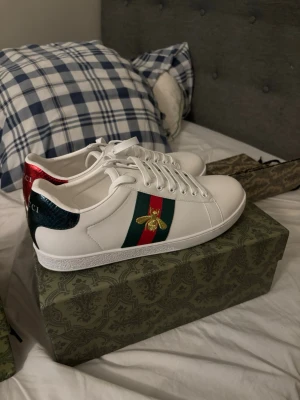 Gucci Ace vita sneakers med bi - Säljer ett par vita Gucci Ace sneakers i läder med klassiska gröna och röda ränder på sidan och en guldbroderad bi. Skorna har vita snören, låg profil och metallic-detaljer i rött och grönt på hälen. Perfekta för dig som vill ha en lyxig och stilren look.