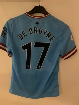 Manchester City matchtröja De Bruyne - Snygg ljusblå Manchester City matchtröja med De Bruyne och nummer 17 på ryggen. Tröjan har klubbmärke, Etihad Airways-tryck och Premier League-logga på ärmen. Detaljer i vinrött och vitt på ärmslut och krage. Perfekt för fotbollsfans!