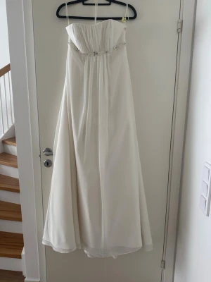 Brudklänning  - Elegant brudklänning med vacker pärldetalj under bysten och dekorativa knappar längs ryggen. Klänningen har ett svepande fall och markerad midja. Längd på bruden 167, under byst 83, byst 87.