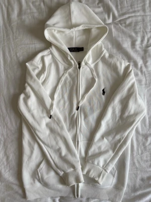Vit hoodie från Polo Ralph Lauren - Snygg vit hoodie med dragkedja från Polo Ralph Lauren. Klassisk design med huva och dragsko, ribbade muddar och fickor framtill. Ikoniska mörkblå loggan broderad på bröstet. Tillverkad i mjukt bomullsmaterial som känns skönt mot huden.