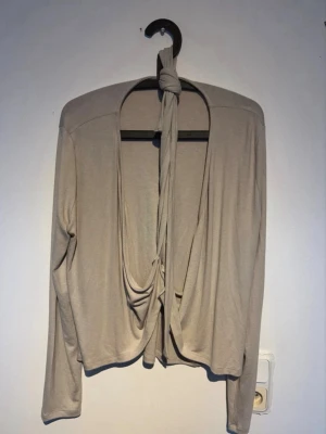 Beige omlottopp med knytband - Trendig beige omlottopp med långa ärmar och knytband framtill. Toppen har en croppad passform och är tillverkad i mjukt, stretchigt material som ger en avslappnad men snygg look. Perfekt att styla över ett linne eller topp. Aldrig använd