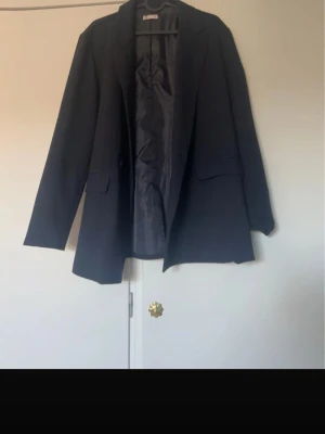 Svart oversized kavaj Gina Tricot - Stilren svart oversized kavaj från Gina Tricot med klassisk krage och två fickor framtill. Kavajen har slät insida och rak passform, perfekt för en trendig och avslappnad look. Materialet är mjukt och följsamt.