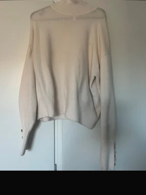 Beige polotröja med pärlknappar - Säljer en beige stickad polotröja med långa ärmar och snygga pärlknappar i guld på ärmsluten. Tröjan har en avslappnad passform och ribbstickade detaljer vid midja och ärmslut. Perfekt för en stilren och trendig look.