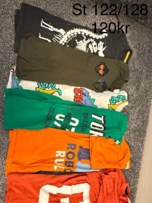 Färgglada t-shirts med tryck, 6-pack - Säljer ett paket med sex t-shirts i olika färger och tryck. Här finns bland annat en grön t-shirt med text, en orange med robot-tryck, en svart med skelettmotiv, en olivgrön med apa, en röd med stor vit bokstav och en vit med färgglada figurer. Perfekt för dig som gillar variation och coola prints.