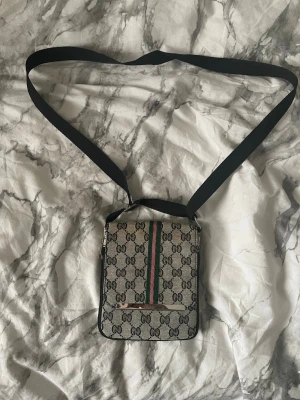 Gucci axelväska med GG-mönster - Snygg axelväska från Gucci i beige och svart med klassiskt GG-mönster och grönt/rött band framtill. Väskan har justerbar svart axelrem och magnetlås. Materialet är canvas med detaljer i läder. Perfekt för dig som vill ha en stilren och ikonisk accessoar.