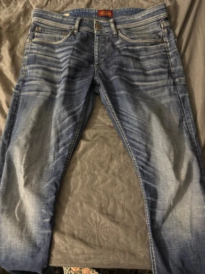 Blå slitna jeans från Jack & Jones - Säljer ett par blå jeans från Jack & Jones med snyggt slitna detaljer och klassisk femficksmodell. Jeansen har en tvättad look med ljusare partier och kontrastsömmar. Perfekta för en avslappnad och trendig stil.