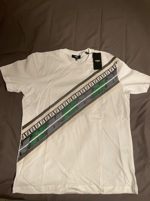 Vit Fendi t-shirt med diagonal logga - Säljer en vit t-shirt från Fendi med diagonal rand i svart, grön och beige samt Fendi-logga längs bröstet. Klassisk rund hals och korta ärmar. Perfekt för dig som gillar exklusiva märken och vill sticka ut med en unik design.