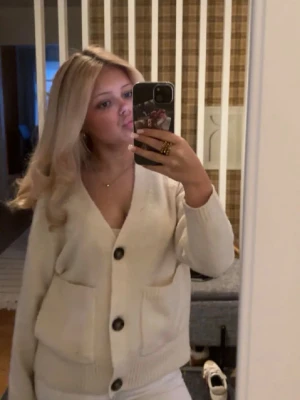 Vit kofta med stora knappar - Mysig vit kofta med v-ringning och tre stora bruna knappar framtill. Koftan har två rymliga fickor och ribbstickade muddar vid ärmslut och nederkant. Perfekt för lager-på-lager och enkel att styla till olika outfits.