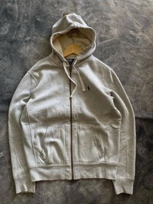Polo Ralph Lauren zip hoodie - Snygg grå hoodie med dragkedja från Polo Ralph Lauren. Klassisk design med huva och dragsko, två fickor framtill och broderad logga på bröstet. Perfekt för en avslappnad och stilren look.