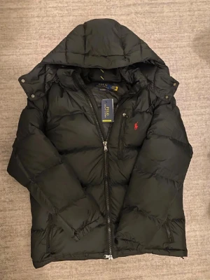 Polo Ralph Lauren Jacka - Snygg svart pufferjacka från Polo Ralph Lauren med huva och dragkedja framtill. Jackan har två sidofickor med dragkedja och den klassiska röda loggan broderad på bröstet. Perfekt för kalla vinterdagar och riktigt skön att bära.