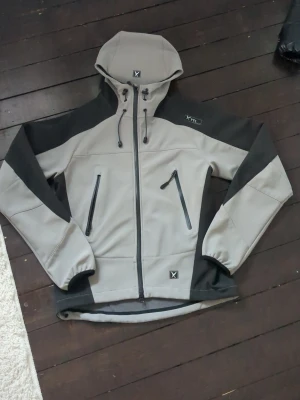 Grå softshelljacka med huva XTC - Snygg grå softshelljacka från TTC med svarta detaljer och dragkedjor. Jackan har två snedställda fickor framtill, justerbar huva med dragsko och elastiska muddar vid ärmsluten. Pefekt för skidåkning,vandring osv vind och vattentät.