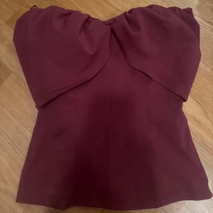 Vinröd axelbandslös topp - Säljer en vinröd axelbandslös topp med snygg drapering över bysten och figurnära passform. Toppen har en trendig och stilren look som passar perfekt till jeans eller kjol. Materialet känns mjukt och stretchigt för en bekväm känsla. Passar s