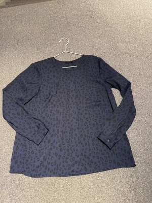 Mörkblå leopardmönstrad blus Lindex - Snygg mörkblå blus från Lindex med svart leopardmönster. Blusen har rund halsringningm och långa ärmar. Knappar fram och vid ärmslut. Tillverkad i ett lätt och mjukt tyg som ger en skön känsla. Perfekt för dig som gillar trendiga mönster och stilren design.