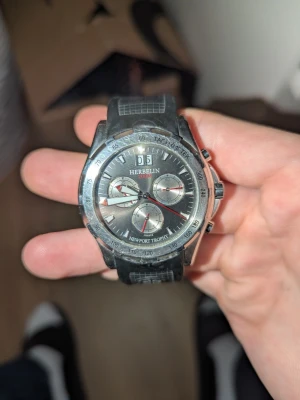 Michel Herbelin Newport Trophy klocka - Snygg kronografklocka från Michel Herbelin, modell Newport Trophy. Klockan har svart urtavla med silverdetaljer och röda visare, samt svart armband med rutmönster. Boetten är i silverfärgad metall och klockan har flera små urtavlor och tachymeter. Köpt för 7000!