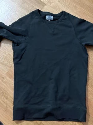 Svart sweatshirt från Crocker XS - En stilren svart sweatshirt från Crocker i storlek XS. Tröjan har rund halsringning och ribbade muddar vid ärmslut och nederkant. Perfekt för en avslappnad och enkel look. Tillverkad i mjukt material som känns skönt mot huden.