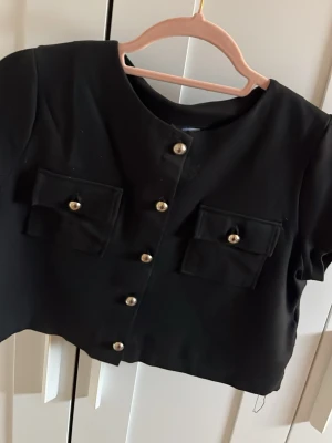 Svart croppad topp med guldknappar - Stilren svart croppad topp med korta ärmar och dekorativa guldknappar framtill. Toppen har två fickor på bröstet med matchande knappar och en rak, enkel passform. Perfekt för att lyfta vilken outfit som helst.