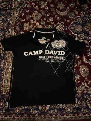 Svart piké Camp David med tryck - Svart pikétröja från Camp David i storlek XL men passar som en Large. Snyggt tryck och brodyr på både fram- och baksida ger en sportig look.