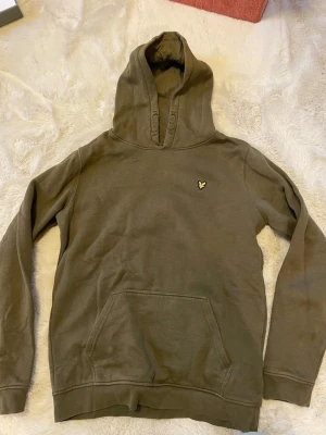 Olivgrön hoodie från Lyle & Scott - Snygg olivgrön hoodie från Lyle & Scott med klassisk gul logga på bröstet. Perfekt för en avslappnad och stilren look. Storleken är 14-15 years vilket passar runt 160- 170 cm. Kontakta vid intresse!!