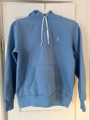 Blå hoodie från Polo Ralph Lauren - Snygg ljusblå hoodie från Polo Ralph Lauren med klassisk gul logga broderad på bröstet. Hoodien har vit dragsko i huvan och en stor känguruficka framtill. Tillverkad i mjukt bomullsmaterial och har ribbade muddar vid ärmslut och nederkant.