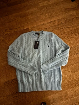 Ljusblå stickad tröja Polo Ralph Lauren - Snygg ljusblå kabelstickad tröja från Polo Ralph Lauren med klassisk rund halsringning och svart broderad logga på bröstet. Tröjan har ribbade muddar vid ärmslut och nederkant. Perfekt för dig som gillar stilrena och tidlösa plagg.