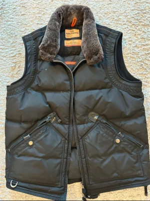 Svart dunväst från Parajumpers - Snygg svart dunväst från Parajumpers Masterpiece Series med fluffig grå fuskpäls på kragen och coola detaljer som dragkedjor och stora fickor framtill. Perfekt för lager på lager och streetwear-vibe.