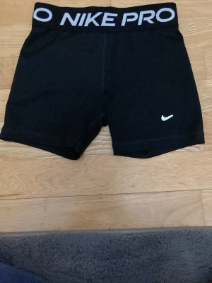 Svarta Nike Pro träningsshorts för barn  - Säljer ett par svarta Nike Pro shorts med bred resår i midjan där det står 'NIKE PRO' i vitt. Tight passform och klassisk Nike-logga på benet. Perfekta för träning och sportiga aktiviteter.