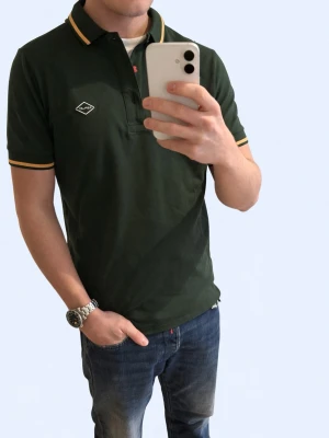 Mörkgrön piké från Replay - Snygg mörkgrön pikétröja från Replay med gul detaljrand på krage och ärmslut. Klassisk passform med knappslå och diskret Replay-logga på bröstet. Perfekt till jeans eller chinos för en clean look.