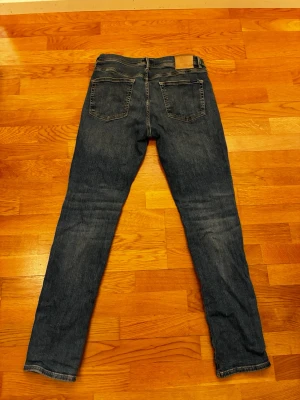 Mörkblå raka jeans - Säljer ett par mörkblå jeans med klassisk femficksdesign och raka ben. Jeansen har en läderpatch bak i midjan och subtila slitningar för en avslappnad look. Perfekta för dig som gillar en tidlös stil.