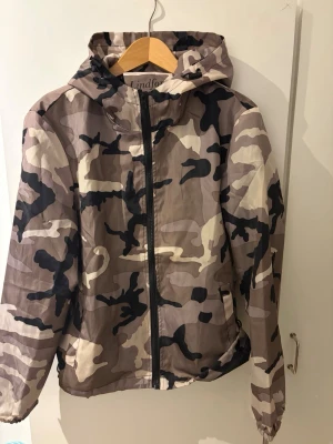 Camouflage vindjacka med huva - Säljer en snygg vindjacka från Lindfors med huva och dragkedja framtill. Jackan har ett fett camouflage-mönster i beige, brun, svart och grå toner. Perfekt för dig som gillar streetstyle och vill sticka ut. Jackan är lätt och har elastiska muddar vid ärmslut och nederkant. Kontakta för mer info! ABS! Pris går att diskutera! 