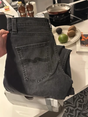 Svarta Nudie Jeans med bakficka - Säljer ett par svarta jeans från Nudie Jeans med klassisk femficksmodell och vågig söm på bakfickan. Jeansen har rak passform och normal midja. Perfekta för dig som gillar stilrena och tidlösa jeans i slitstark denim.