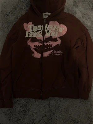 Brun hoodie Legacy Studios S - Brun hoodie från Legacy Studios i 100% ekologisk bomull. Dragkedja framtill, huva och känguruficka. Tryck med texten 'Thanks for being you' och rosa grafiskt motiv med läppar på bröstet. Perfekt för en chill och avslappnad stil.