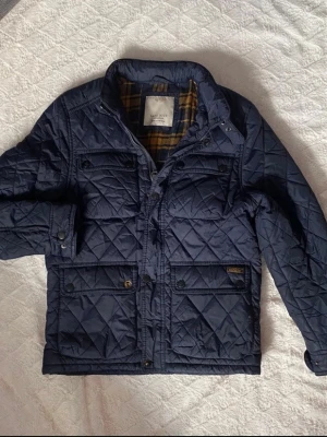 Mörkblå quiltad jacka Zara 152cm - Snygg mörkblå quiltad jacka från Zara med rutigt innerfoder i gult och svart. Jackan har fyra framfickor med knappar, hög krage med tryckknapp och detaljerade sömmar. Perfekt för kalla dagar och har en klassisk, tidlös look.