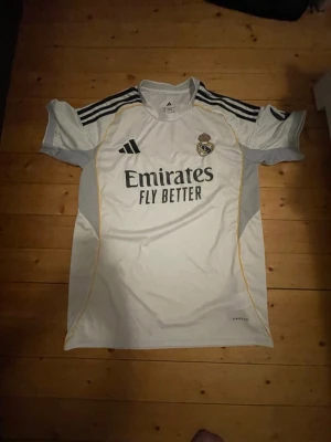Real Madrid matchtröja Adidas vit - Säljer en vit Real Madrid matchtröja från Adidas med svarta detaljer och gula linjer. Tröjan har klubbmärke på bröstet, Adidas-logga och texten 'Emirates Fly Better' på framsidan. Tillverkad i lätt och ventilerande material, perfekt för fotbollsträning.