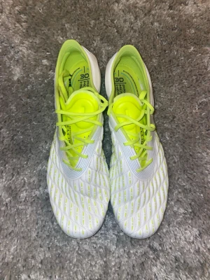 Vita och neongula under armour fotbollsskor - Säljer ett par snygga fotbollsskor från under Armour i vitt och neongult. Skorna har en modern design med struktur på ovandelen för bättre bollkontroll och snörning framtill. Tillverkade i syntetmaterial med greppvänliga dobbar för spel på gräs.